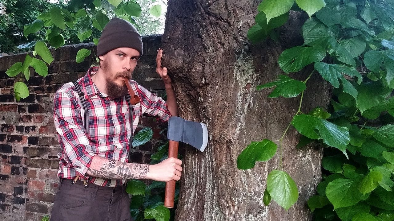 Carlos Eriksson dressed as a lumbersexual and posing next to a tree holding a faxe card board axe.
